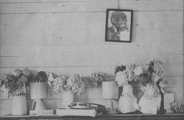 wall with crooked picture of man praying, shelf with vases(?) of flowers